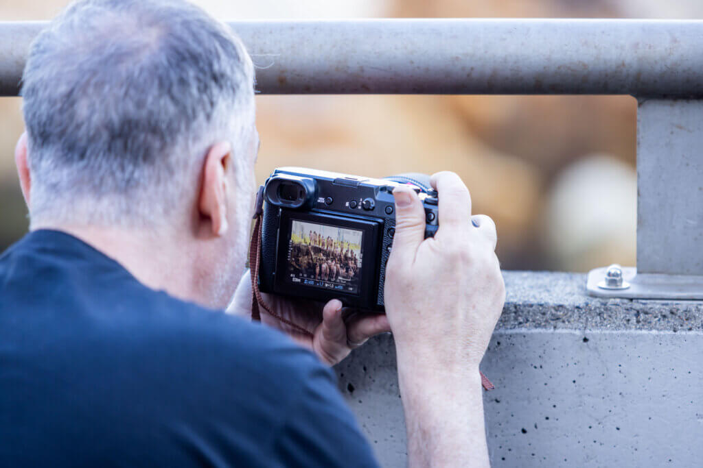 A man with short gray hair and a black shirt is using a DSLR camera to take photos. Positioned close to a concrete railing, he appears deep in concentration as the camera's display screen shows a preview of an outdoor scene. The background is blurred, hinting at his role as an ambassador for Melt Festival 2024.