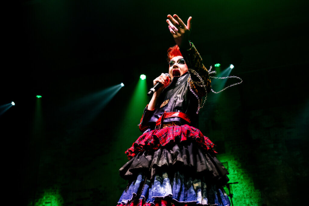 A performer in vibrant, elaborate clothing, featuring a layered skirt and a studded leather jacket, sings passionately on stage. The background is dimly lit with green spotlight effects, intensifying the dramatic atmosphere of the performance.