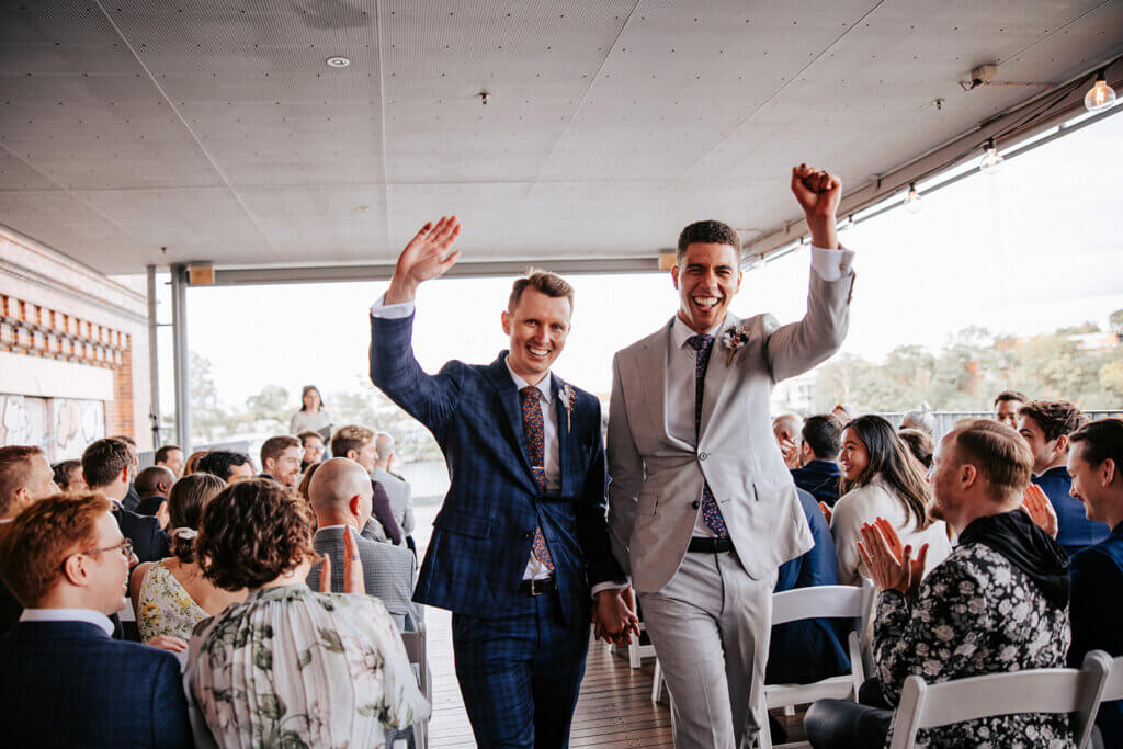 A joyful couple, dressed in suits, walk hand-in-hand down an aisle at their outdoor wedding ceremony while guests applaud and cheer. One person in a blue checkered suit waves, and the other in a light gray suit raises a fist in celebration.