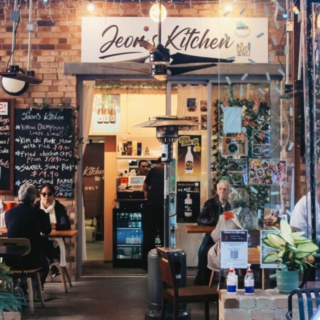 A cozy restaurant entrance named "Jeon's Kitchen" with brick walls and a wooden sign. Diners sit at tables outside under string lights. The interior is warmly lit with a display of photos and a menu board. A large, black ceiling fan is mounted above the tables.