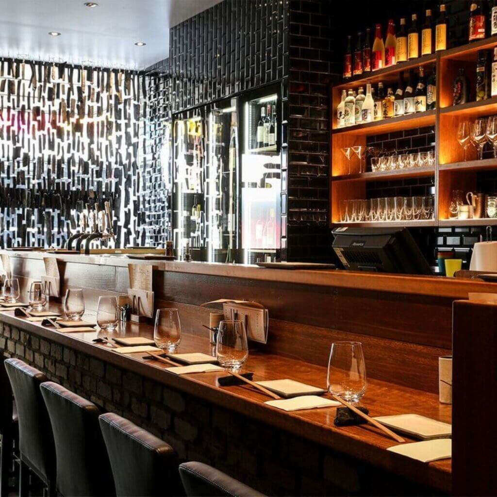 A modern bar with a dim, cozy atmosphere. There are high stools along a wooden bar counter with wine glasses and menus neatly placed. The back wall features shelves lined with bottles and glasses, while a metal decorative screen filters light into the space.