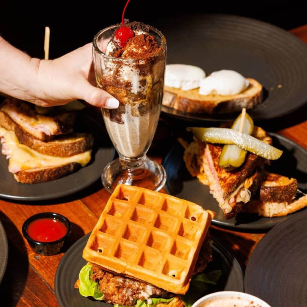 A hand holding a glass with whipped cream and a cherry on top, surrounded by various dishes. The table features a waffle sandwich, pickles on a sandwich, poached eggs on toast, and a plate with sandwich halves, all on dark plates with a wooden table underneath.