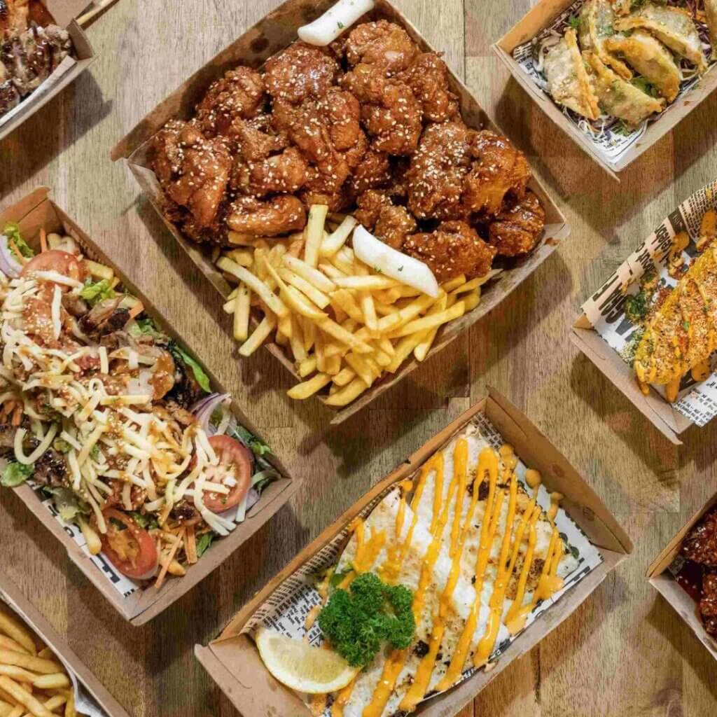 A variety of takeout food items arranged on a table, including a box of crispy fried chicken with sesame seeds and fries, a container of salad with shredded cheese and tomatoes, sushi rolls, and other dishes drizzled with sauces.