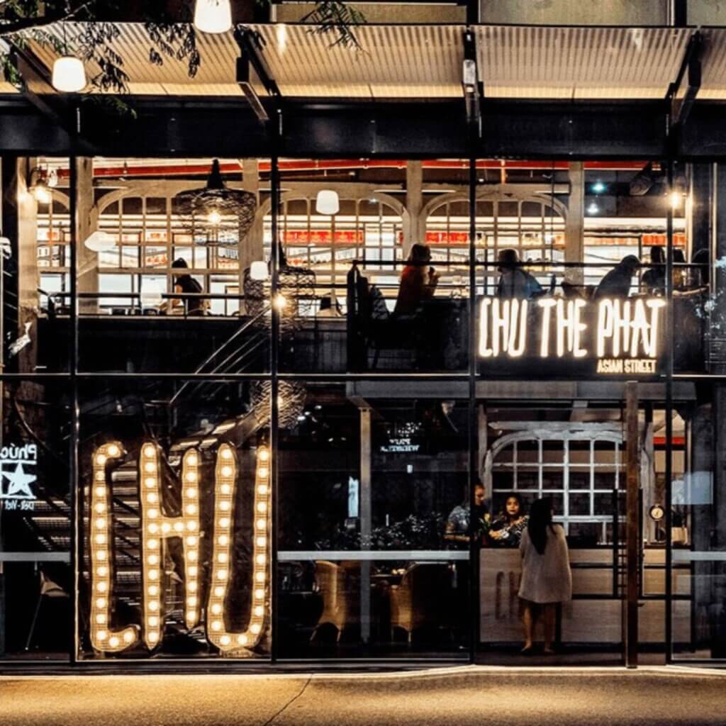 A brightly lit, modern restaurant with large glass windows showcasing its interior. The sign "CHU THE PHAT" is illuminated on the front, and there are patrons dining inside. The entrance features the letters "CHU" in large, marquee lights.