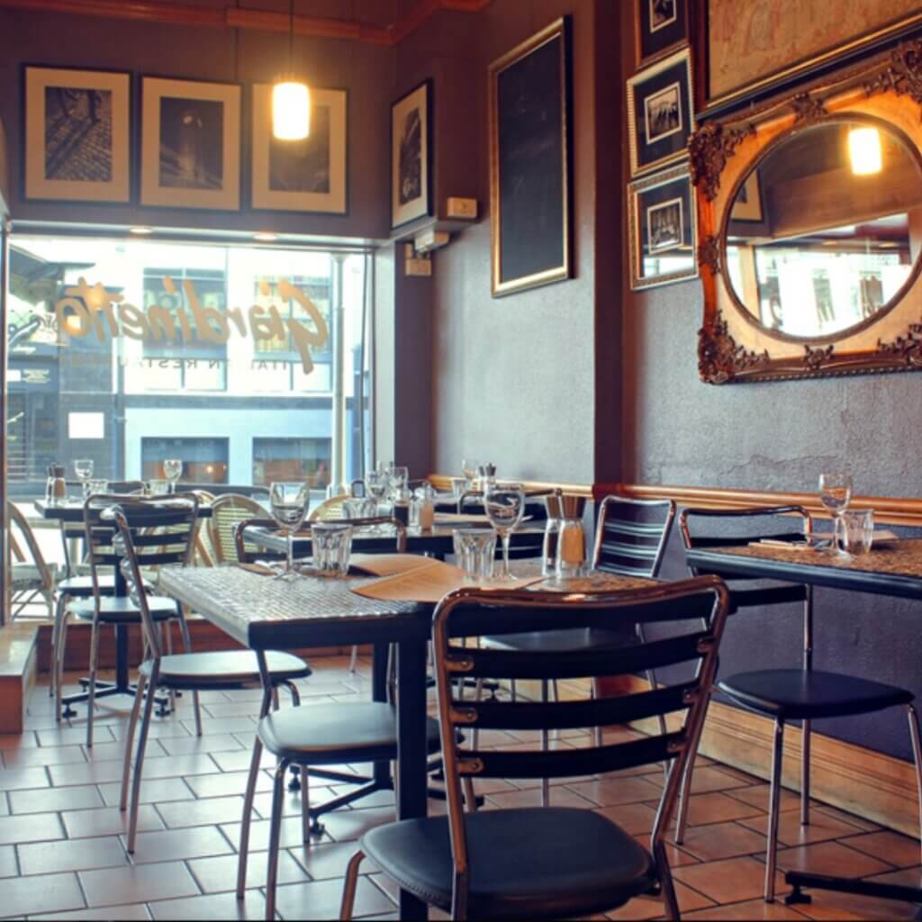 A cozy restaurant interior with neatly arranged tables and chairs. Wine glasses and cutlery are set on the tables. The walls are adorned with framed photos and a large ornate mirror. Soft lighting creates a warm atmosphere. A window shows a glimpse of the street outside.