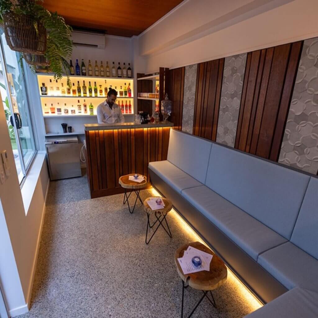 A cozy cafe features a barista standing behind the counter, with shelves stocked with various bottles. There's a long bench with cushions along the wall, small round tables with cups on them, and ambient lighting throughout the space, including under the seating area.
