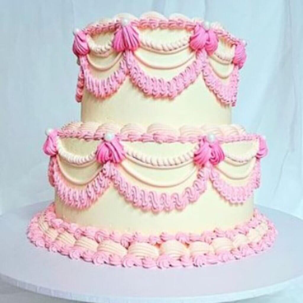 A two-tiered cake with off-white frosting is decorated with intricate pink piping, creating elegant swags and rosettes. The cake sits on a round, white base against a plain, light background.