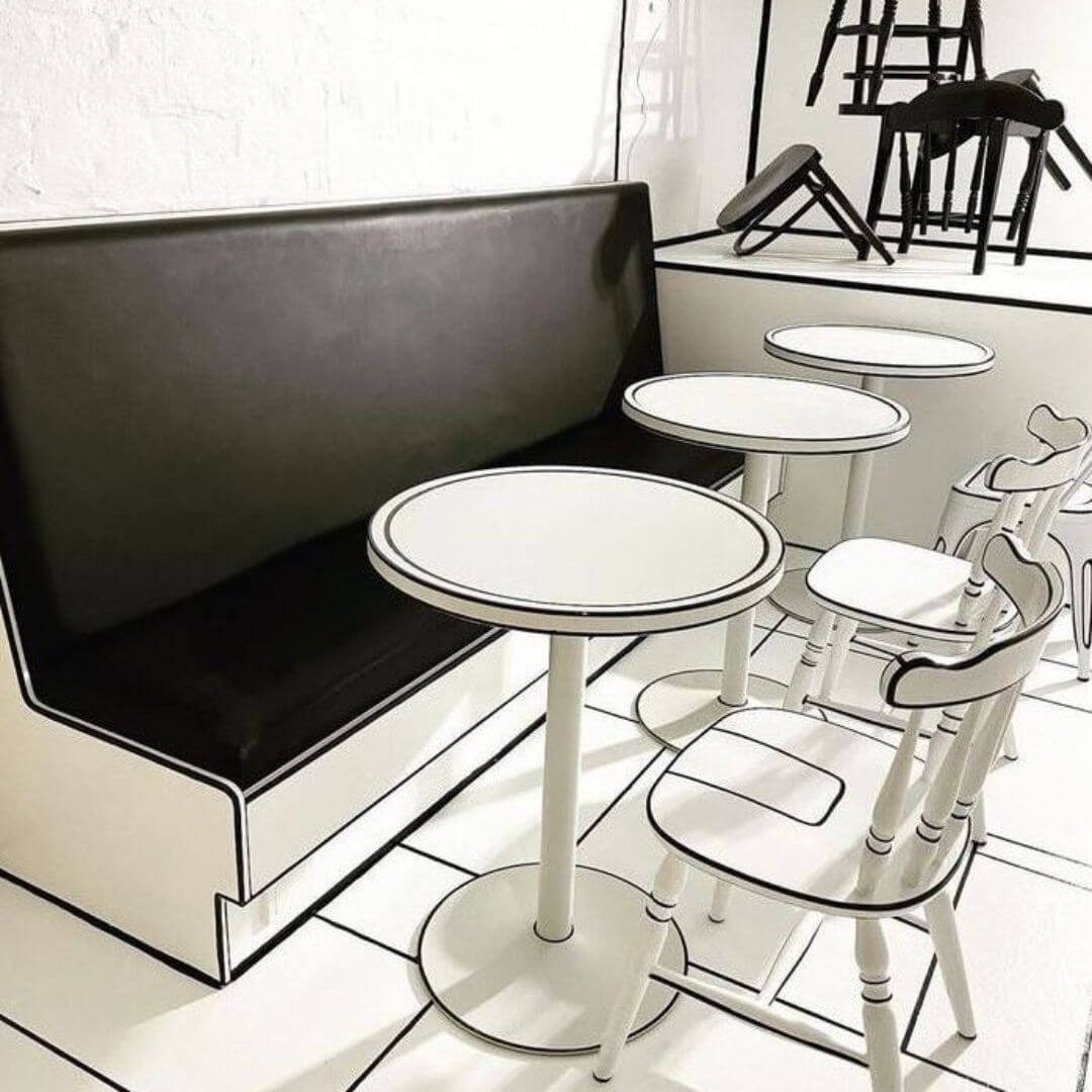 A cafe interior designed to look like a black and white comic book. The furniture, including a black cushioned bench, white tables, and chairs, features bold black outlines. The walls and floor have a similar illustrated style, enhancing the cartoonish effect.