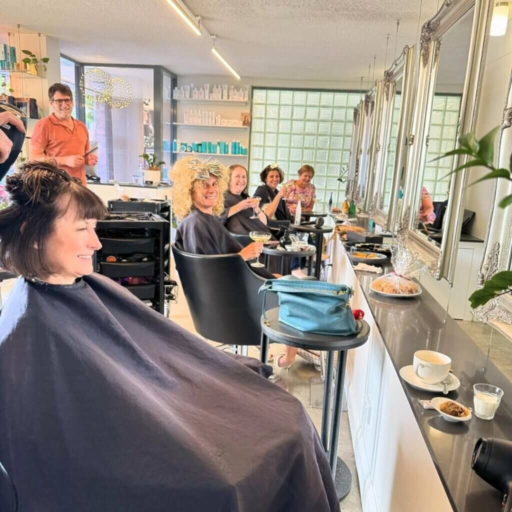 A cheerful group of people at a hair salon is getting their hair done, chatting and smiling in anticipation of Melt Festival 2024. Coffee cups and a purse rest on the counter, while large mirrors line the walls, reflecting the bright, modern interior reminiscent of style paradise.