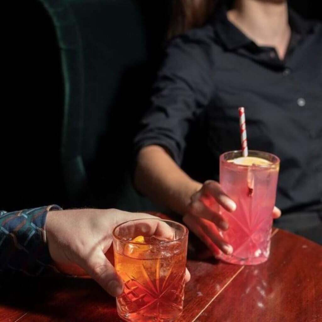 Two people are seated and holding drinks; one has a clear glass with an amber-colored drink and an orange garnish, and the other has a clear glass with a pink drink, a slice of lemon, and a striped straw. The setting appears to be a dimly lit bar.