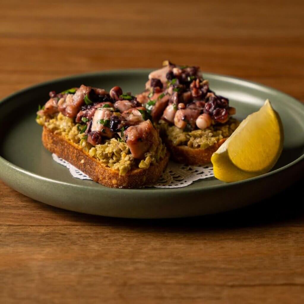 A green plate has two pieces of toasted bread topped with a mixture of diced octopus, herbs, and spices. The bread appears to have an olive tapenade base. A lemon wedge rests on the plate, which is set on a wooden table.