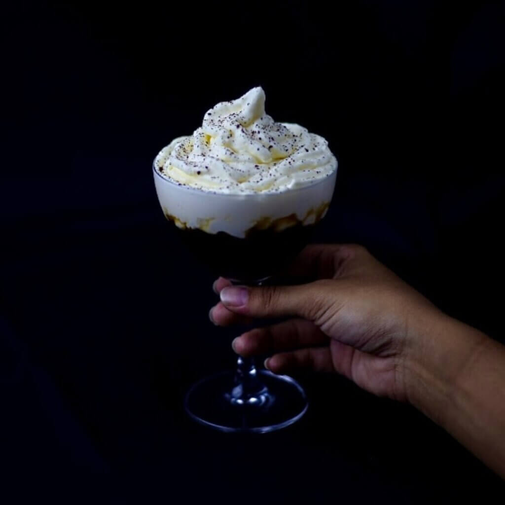 A hand holding a glass filled with dark liquid, topped with a dollop of whipped cream and sprinkled with black specks, evokes the spirit of Paradise Pass against a dark background.