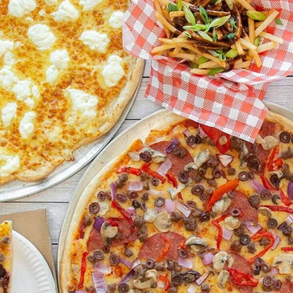 A top view of a table with food, including a cheese pizza, a meat and vegetable pizza topped with olives, red peppers, onions, and mushrooms, and a basket of fries garnished with green onions in a red and white checkered paper.