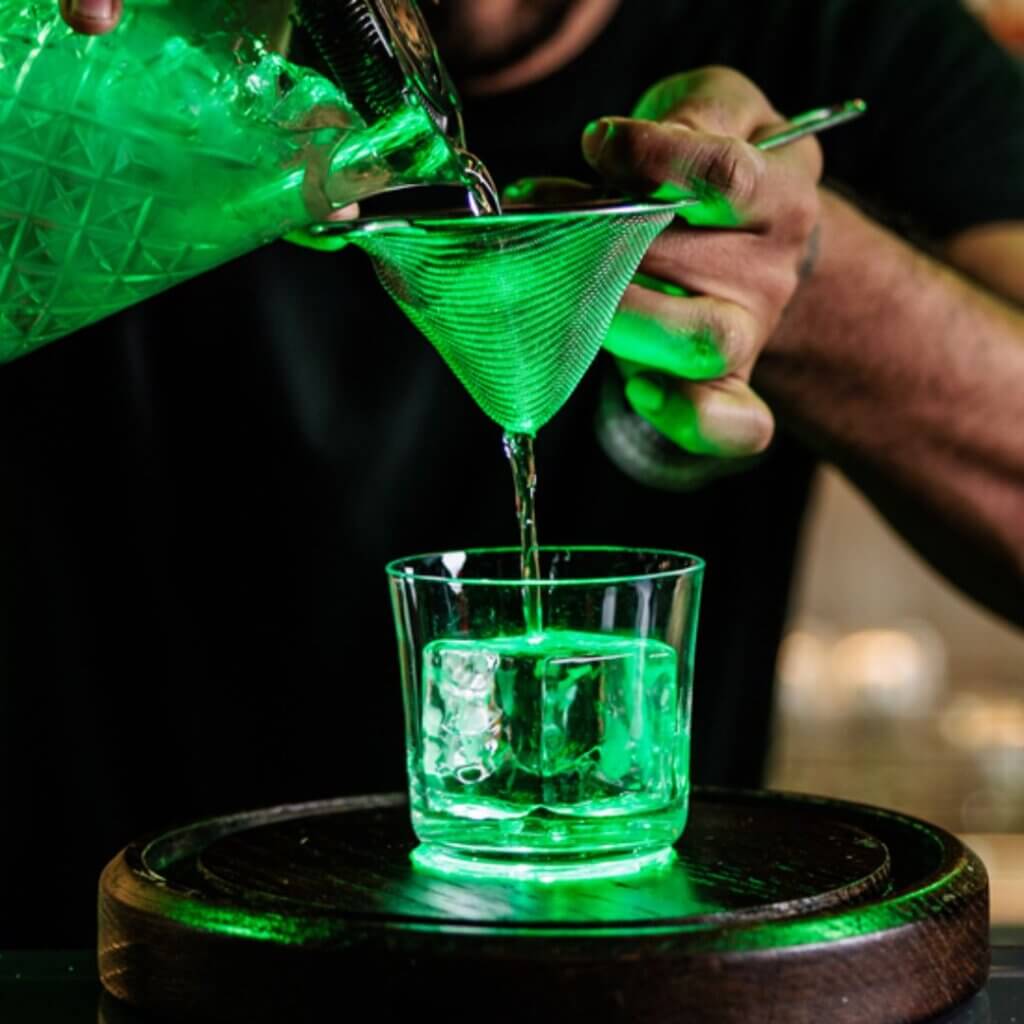 A bartender pours a green cocktail through a mesh strainer into a glass filled with ice cubes. The scene is illuminated by a green light, giving the drink an otherworldly glow. The glass is placed on a wooden surface.