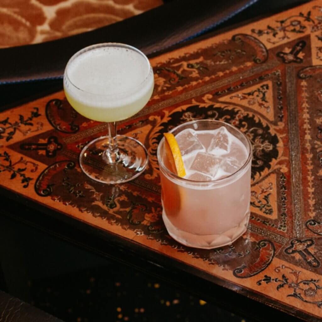 A photo of two cocktails resting on an ornate, engraved wooden table. The drink on the left is a pale green liquid in a coupe glass, while the drink on the right is pink with ice cubes and an orange wedge garnish in a rocks glass.