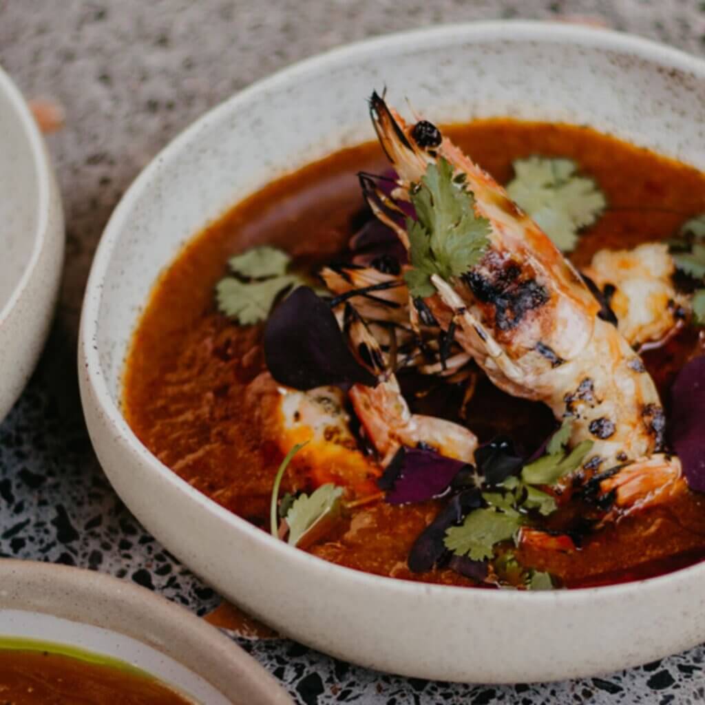 A close-up of a bowl of seafood stew garnished with a large grilled shrimp, cilantro, and purple leaves. The stew is rich in color and appears to be seasoned with various spices. The bowl is set on a textured, stone-like surface.