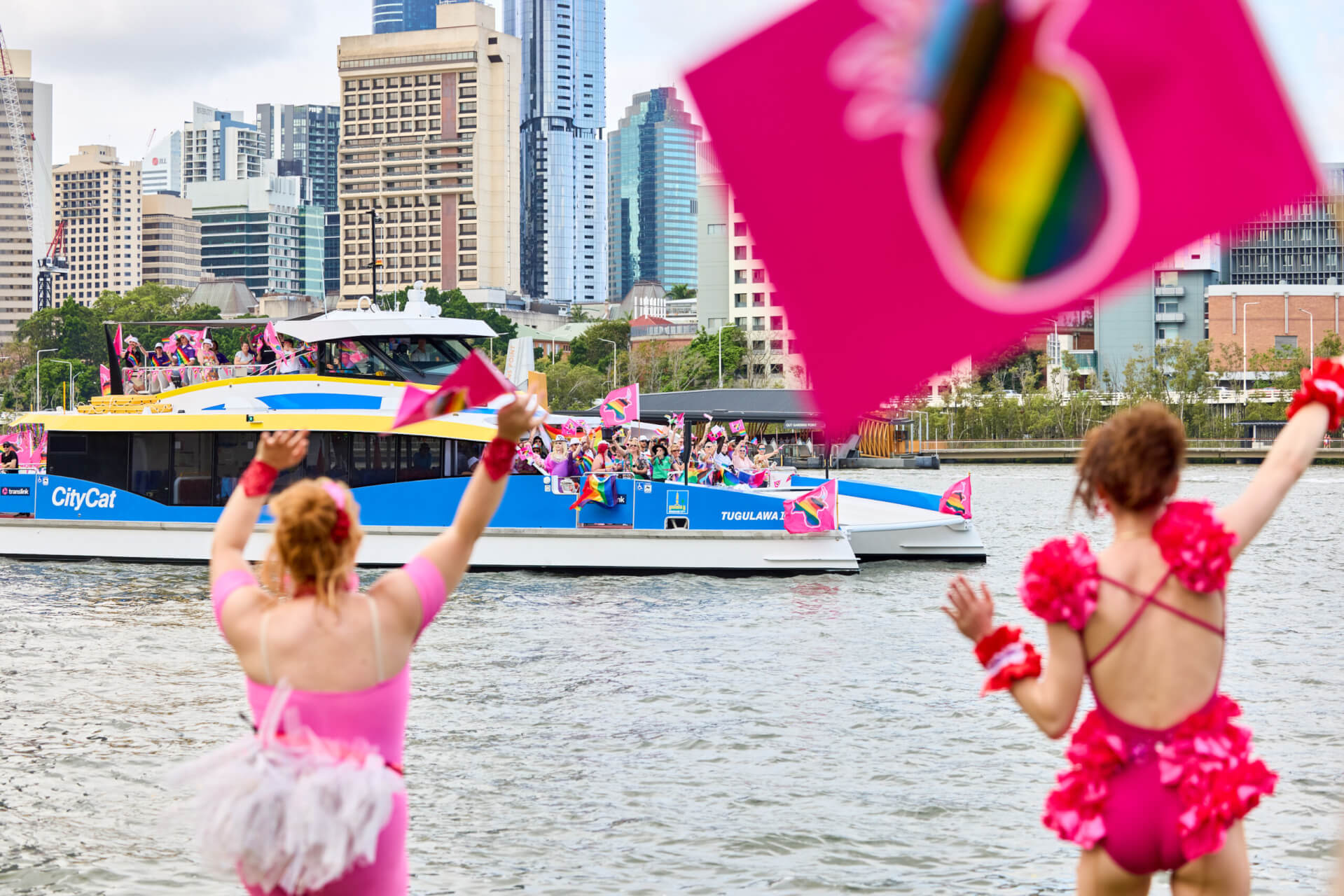 Two people in pink outfits wave rainbow flags as a ferry decorated with rainbow flags passes by on a river. The city skyline is visible in the background, with multiple high-rise buildings.