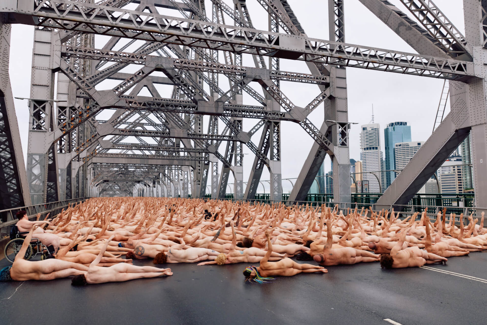 Large group of nude people lying on their backs on a bridge, with arms extended upward. The bridges metal structure frames the scene, and a city skyline is visible in the background.