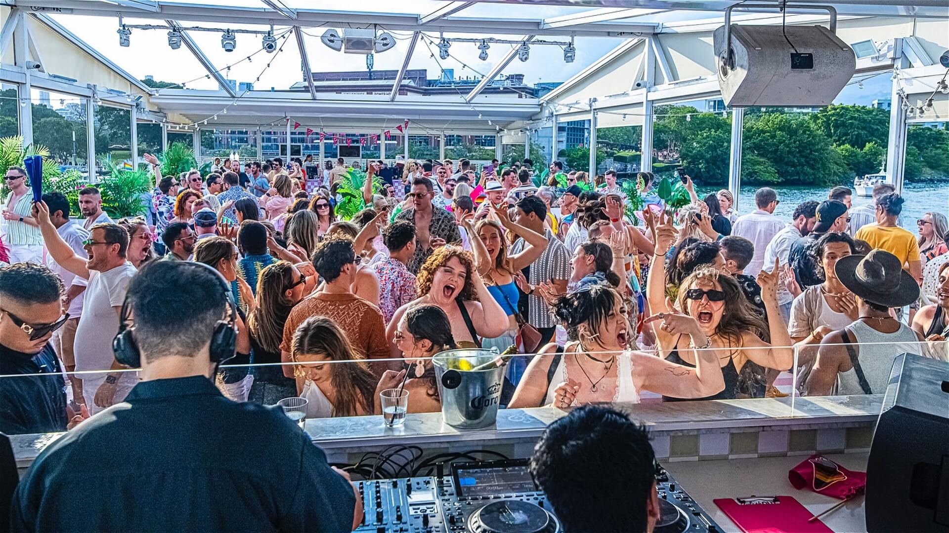 A lively outdoor party with a large crowd dancing and enjoying music from a DJ at a rooftop bar. People are smiling, cheering, and wearing summer clothes under a bright canopy by the water.