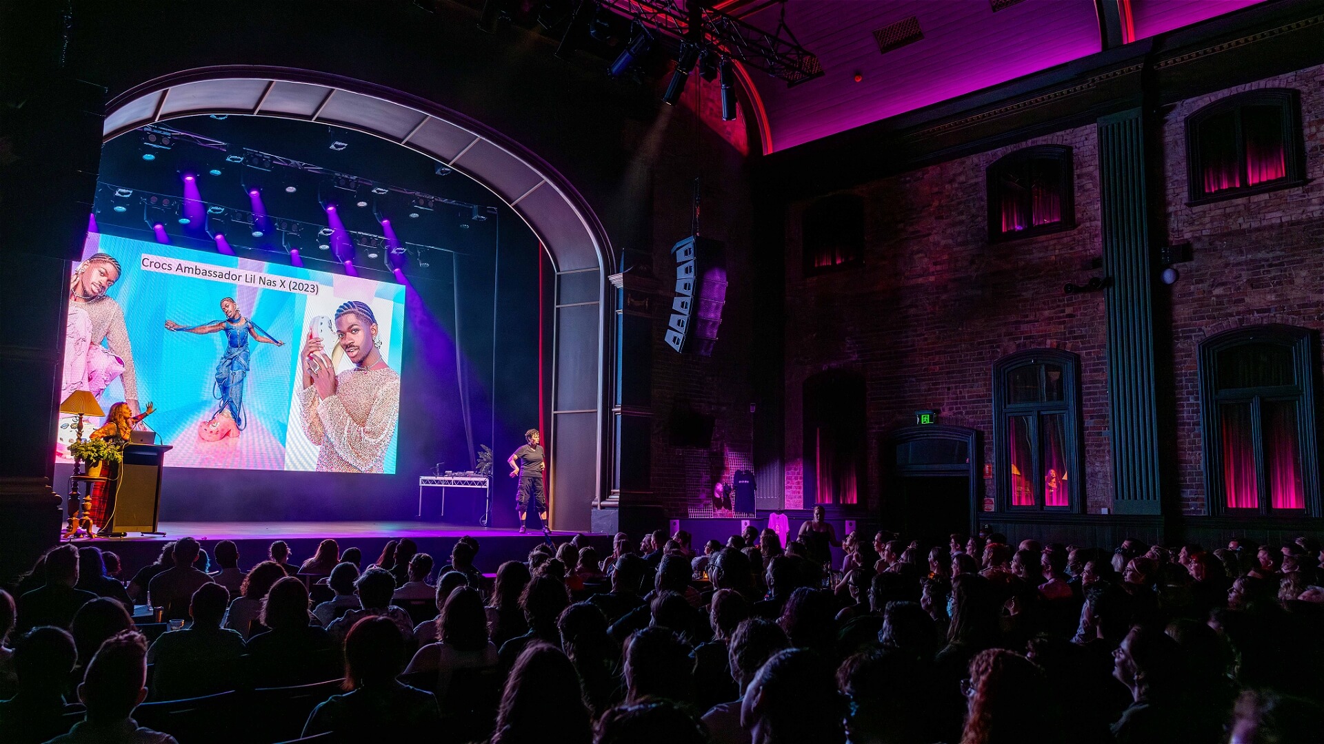 A speaker stands on stage in a theater with purple lighting, presenting to a seated audience. A large screen shows images of people in colorful outfits and the text Cross Ambassadors 14 May 2023.