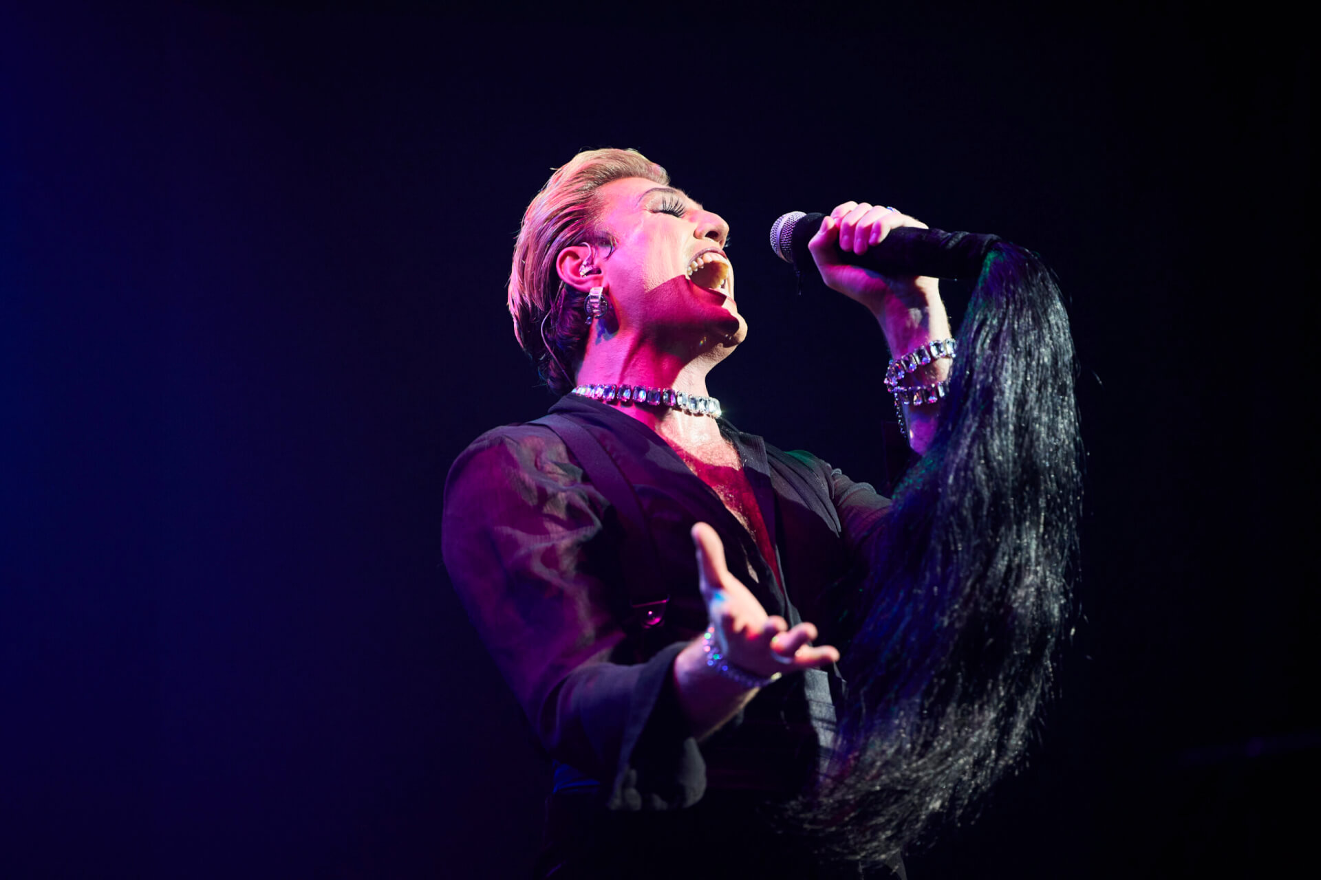 A performer with short blond hair, dramatic makeup, and jewelry passionately sings into a microphone on stage, wearing a sheer black outfit and holding a prop resembling a long black ponytail.