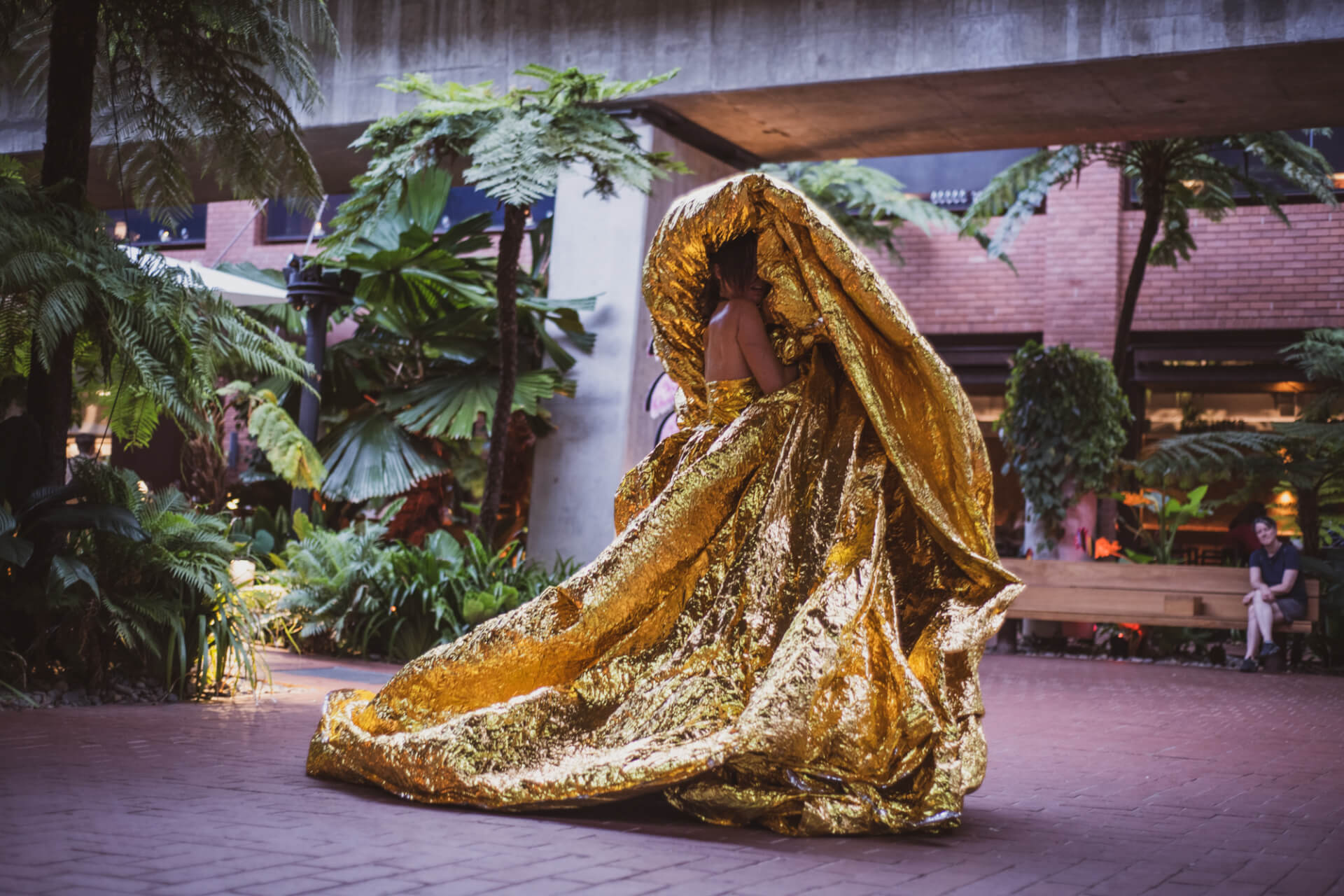 A person outdoors is wrapped in and partially hidden by a large, shiny gold metallic fabric, surrounded by lush green plants at Brisbane’s Melt Festival, with brick buildings and benches in the background.