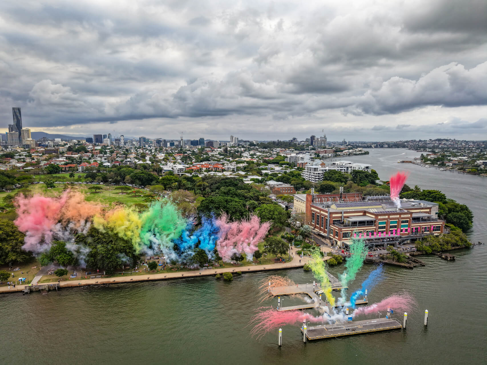 A riverside city scene with colorful smoke bursts in pink, green, blue, and yellow along a walkway and near a large building, under a cloudy sky. The city skyline and water are visible in the background.