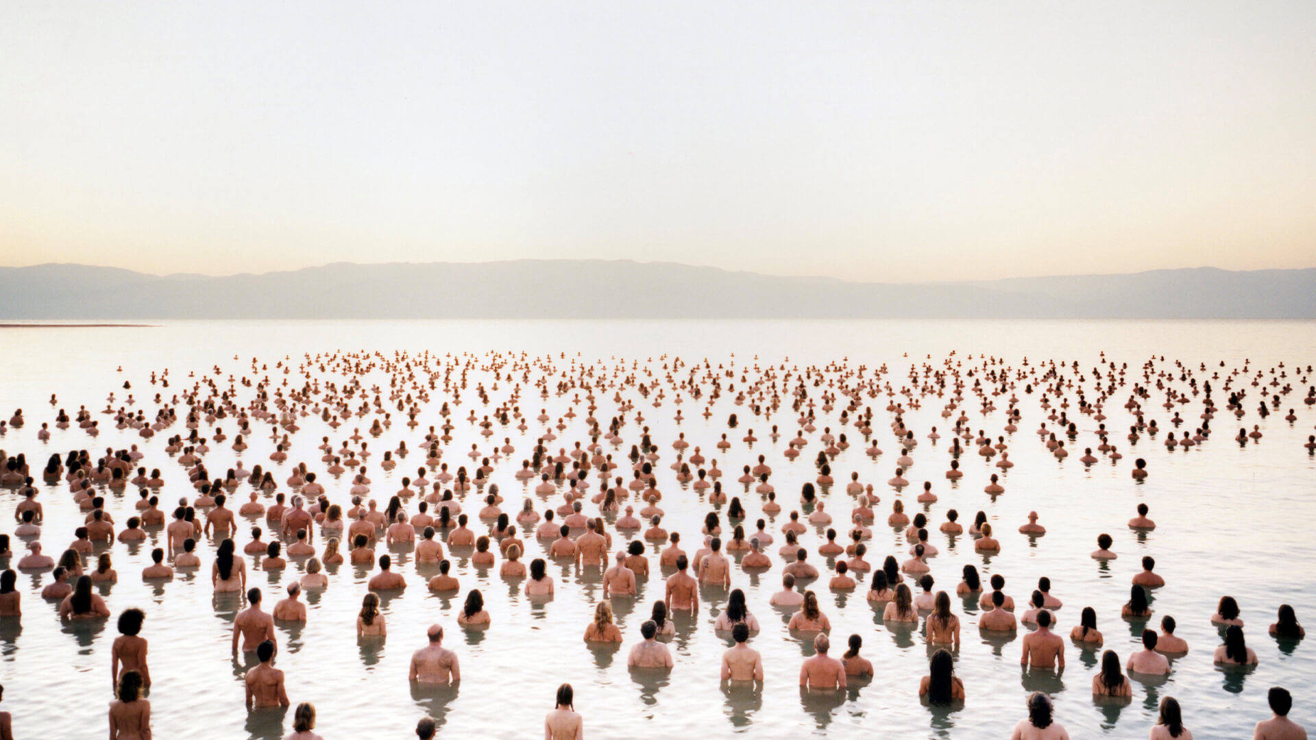 Hundreds of people stand closely packed in shallow water, facing towards the horizon under a pale sky, with distant mountains visible in the background.