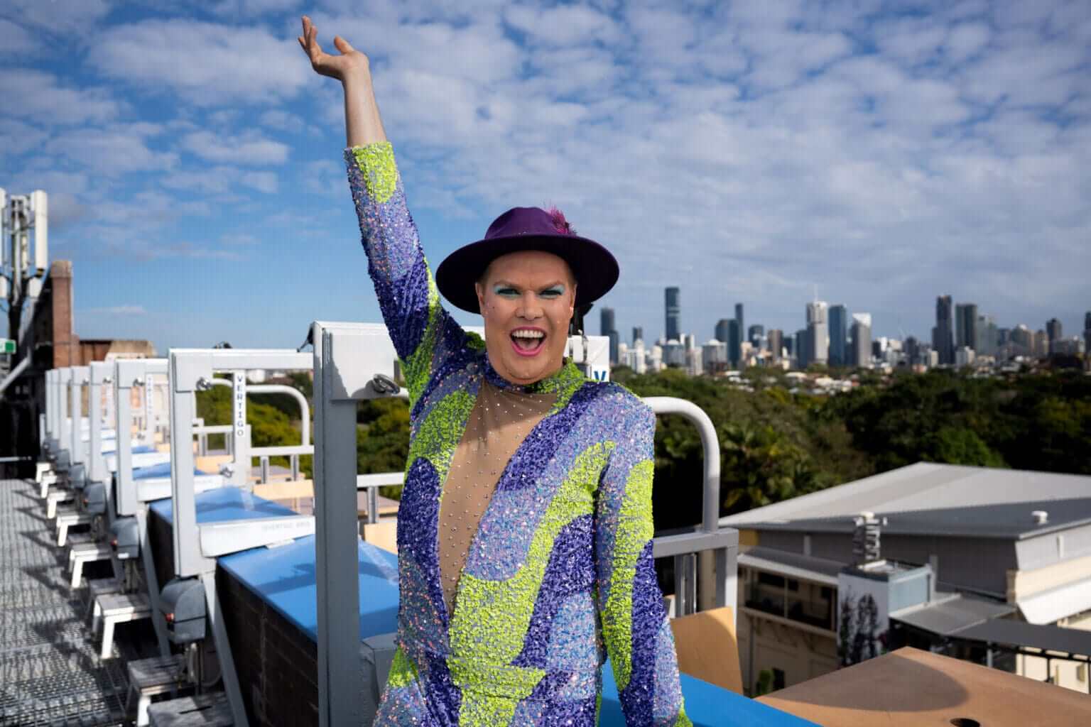 A person in a colorful, sequined outfit and purple hat poses with one arm raised on a rooftop, smiling brightly with a city skyline and blue sky with clouds in the background.