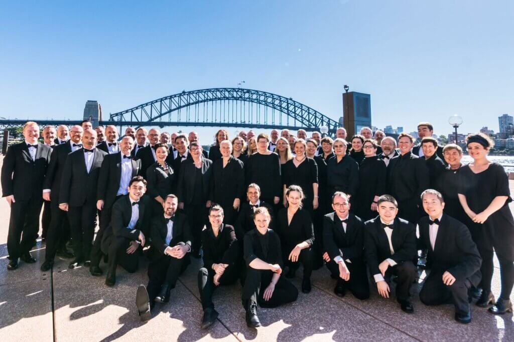 A large group of people dressed in formal black attire pose together outdoors, with the Sydney Harbour Bridge and city skyline visible in the background on a sunny day.