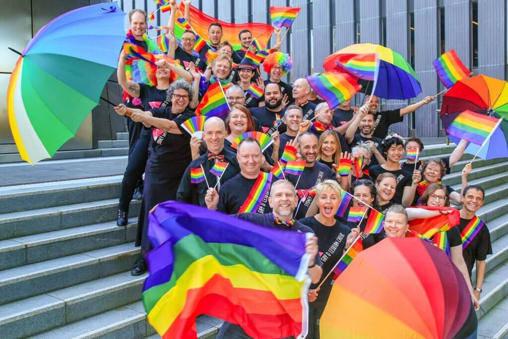A group of people stand and sit on steps outside, smiling and holding rainbow flags and umbrellas. They wear black clothes with rainbow accessories, celebrating LGBTQ+ pride.