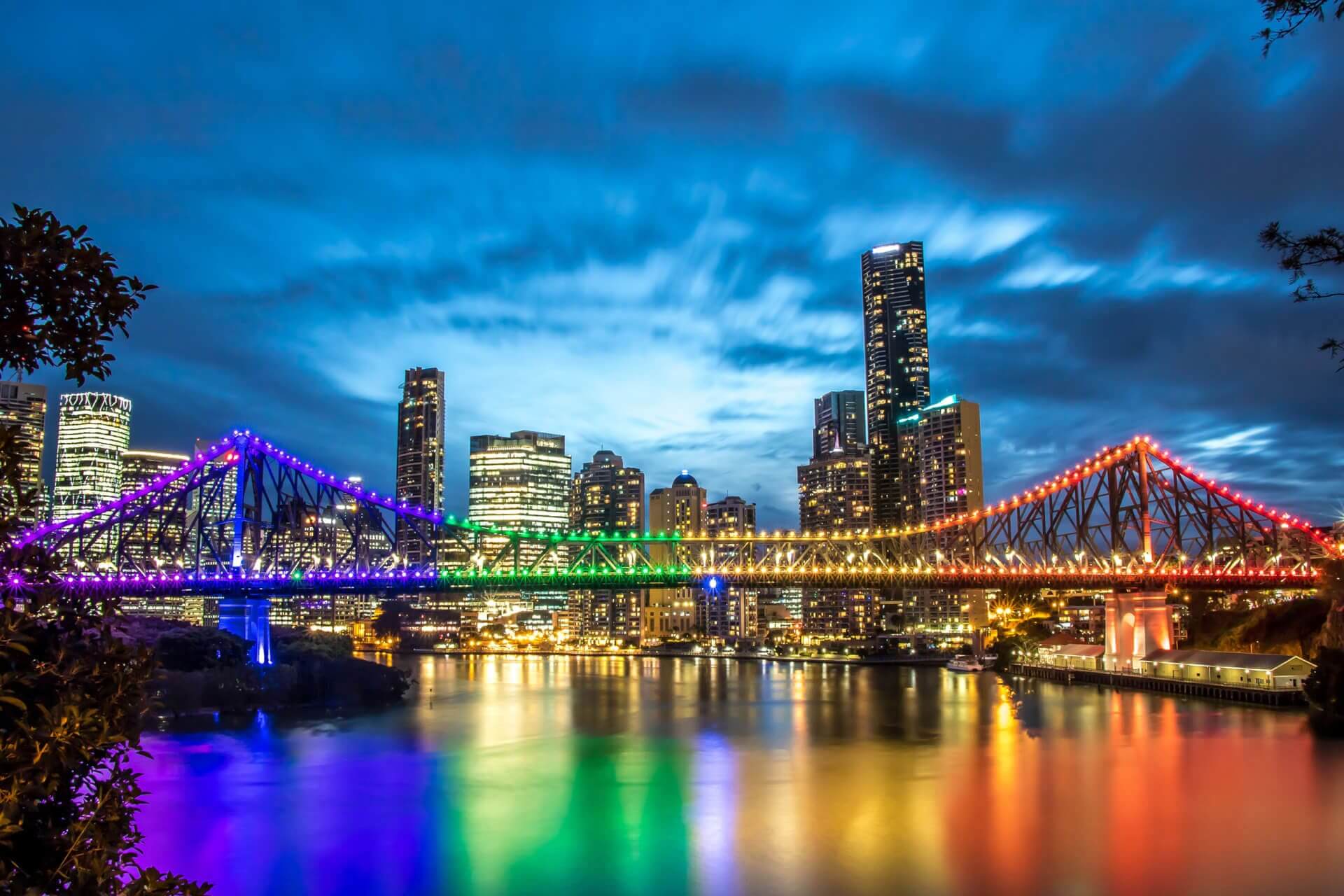 A city skyline at dusk with a brightly lit bridge displaying rainbow colors that reflect on the water below. Tall buildings with illuminated windows rise in the background under a dramatic, cloudy sky.