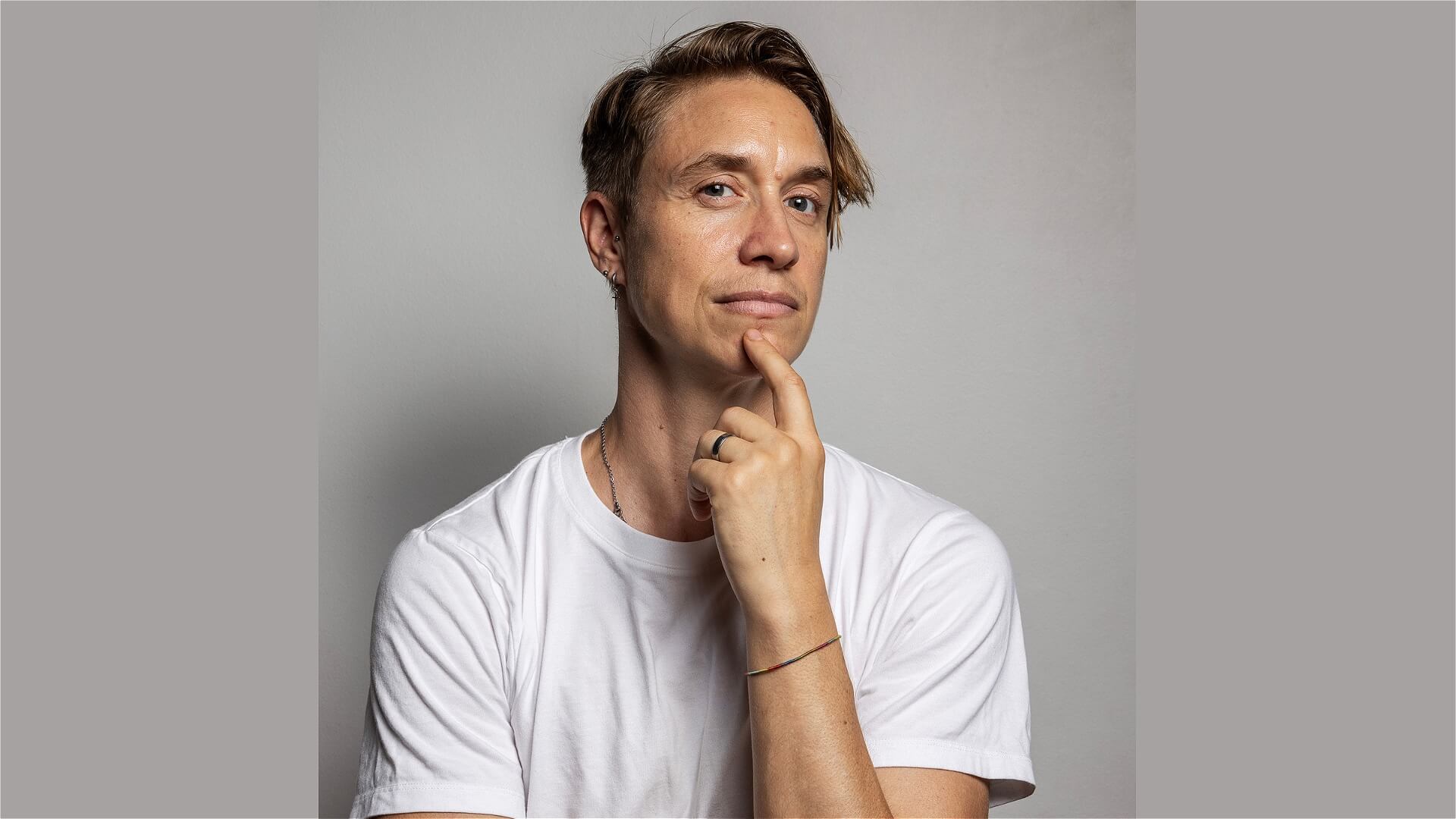 A person with short, light brown hair wears a white t-shirt and gazes thoughtfully at the camera, resting their chin on their hand. The background is plain and gray.