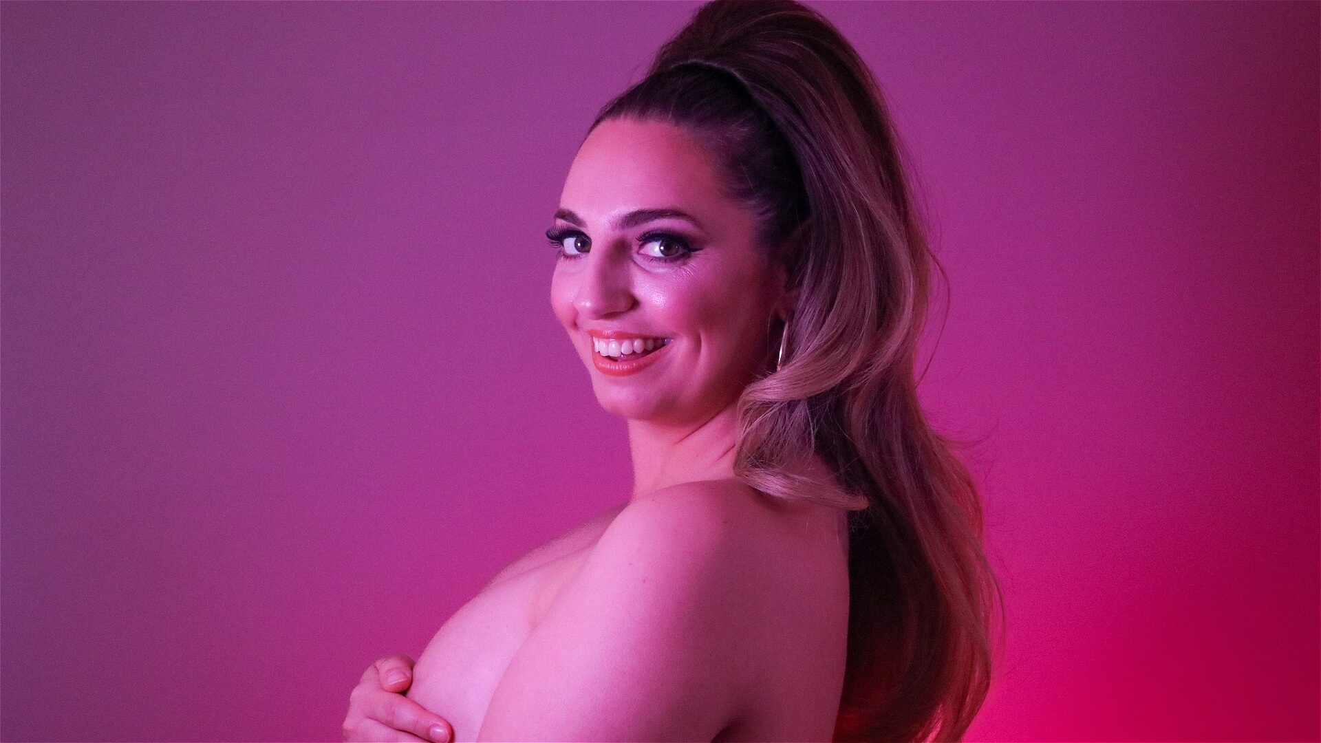 A person with long, styled hair smiles at the camera in front of a pink and purple-lit background, covering their chest with one arm.