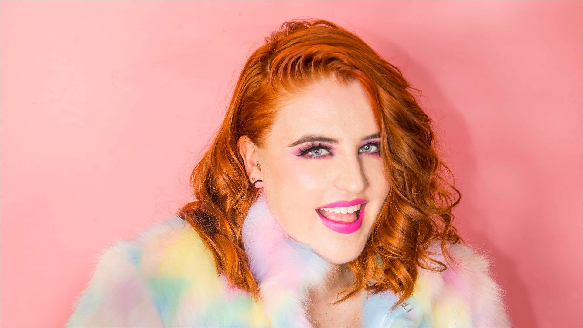 A woman with wavy red hair smiles against a pink background. She wears bright makeup and a pastel-colored faux fur jacket, creating a vibrant, cheerful look.