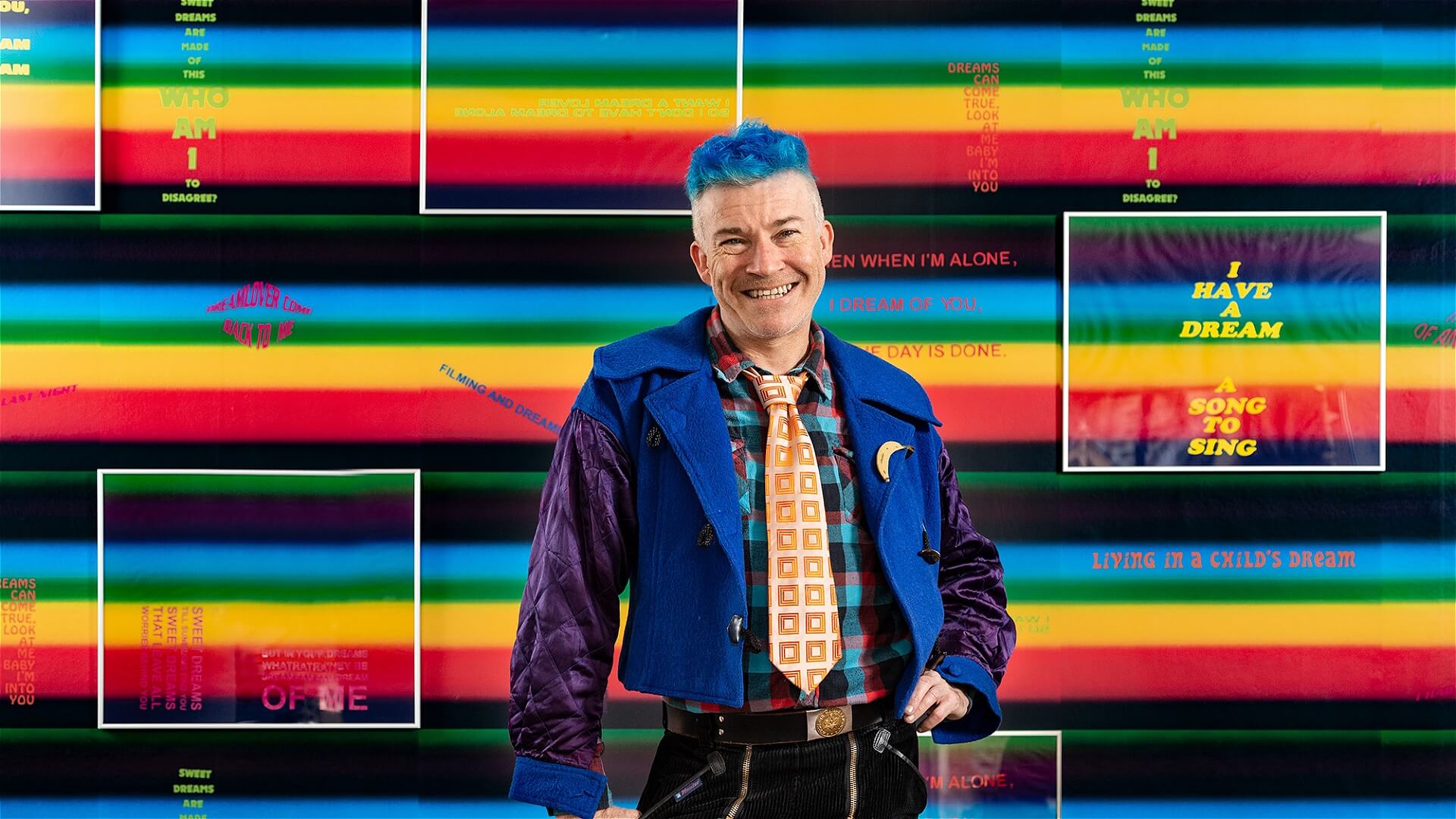 A smiling person with bright blue hair stands in front of a colorful, striped background featuring text, geometric shapes, and various dream-related phrases. They wear a blue jacket, patterned tie, and vibrant clothing.