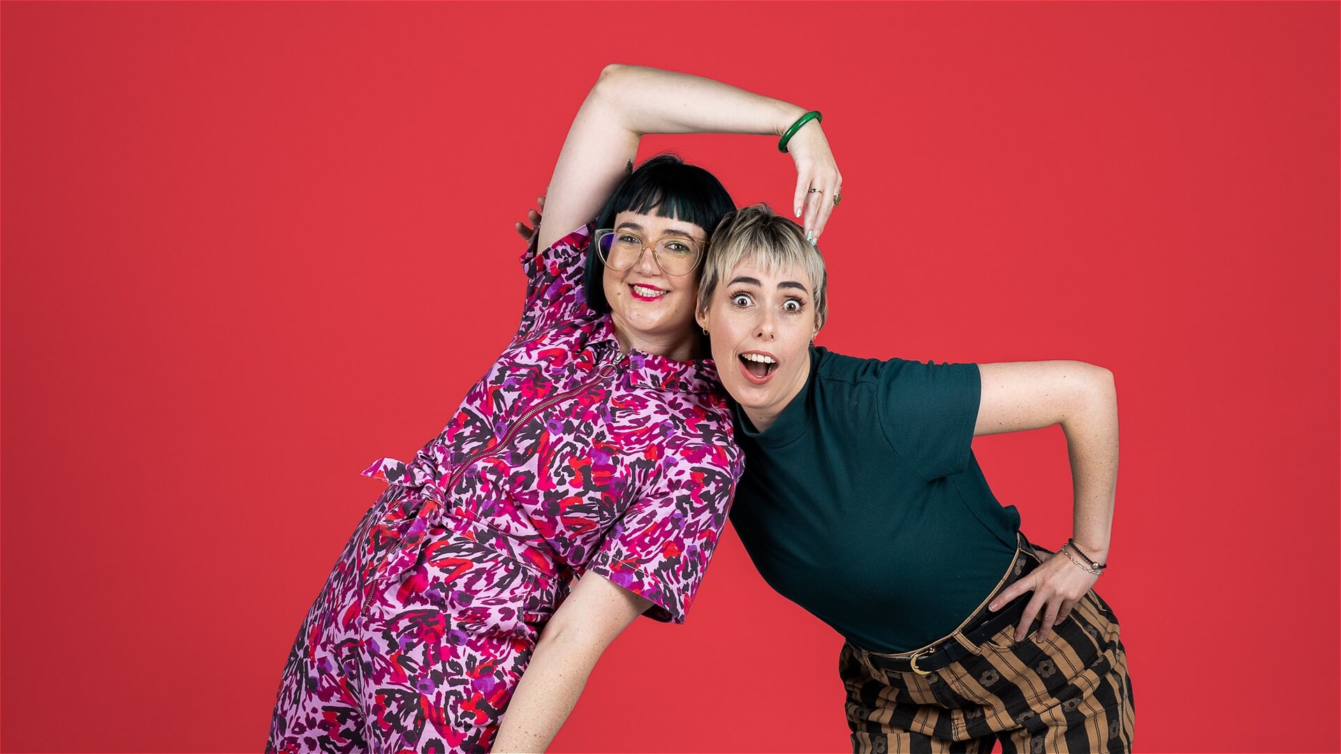 Two people smiling and posing playfully against a bright red background. One wears a colorful pink dress and glasses; the other wears a green t-shirt and striped pants. They lean toward each other with raised arms in a fun, dynamic pose.
