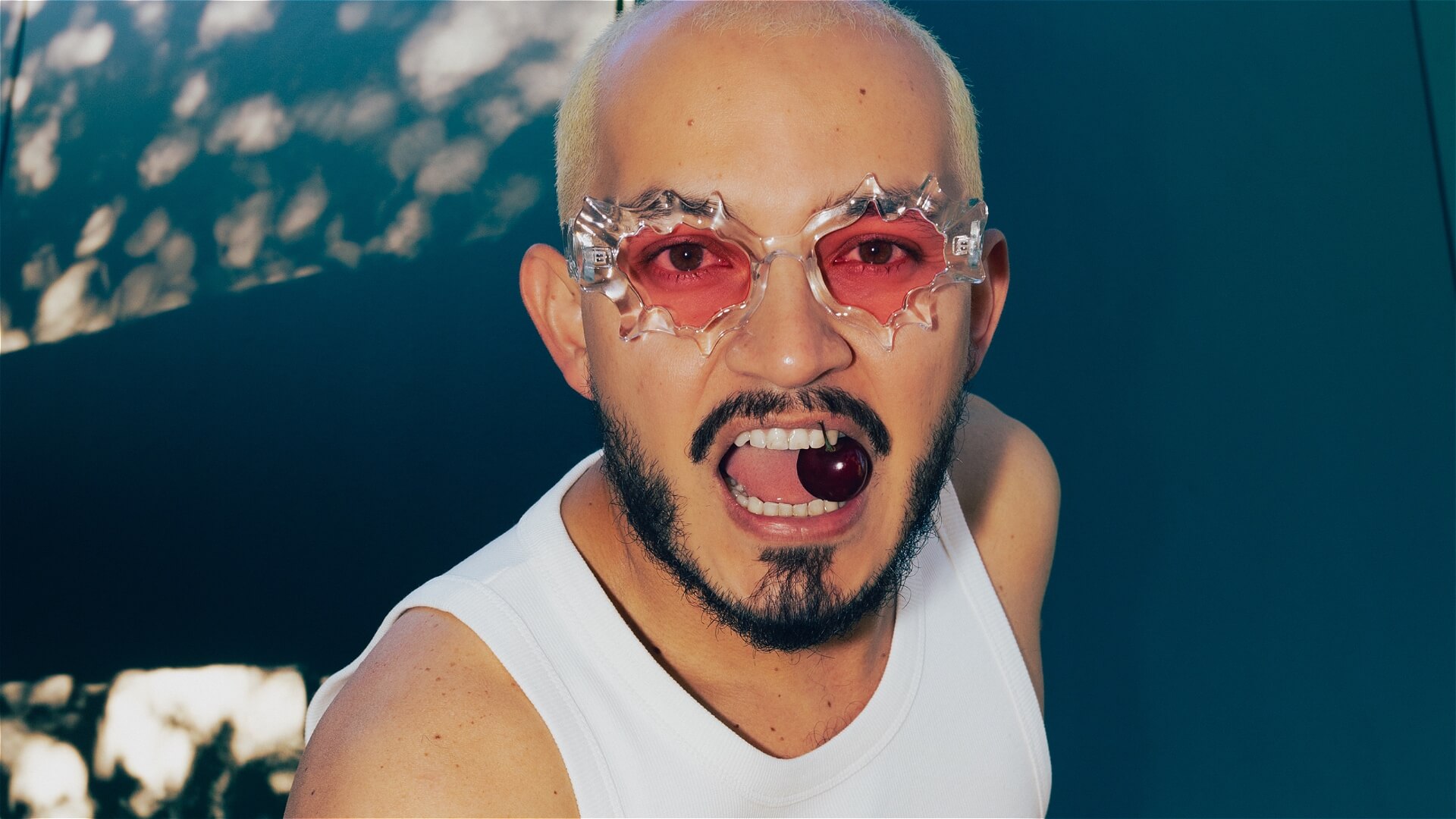 A person with bleached hair and a beard wears spiky, clear-framed glasses with red lenses, a white tank top, and holds a cherry between their teeth, looking into the camera.