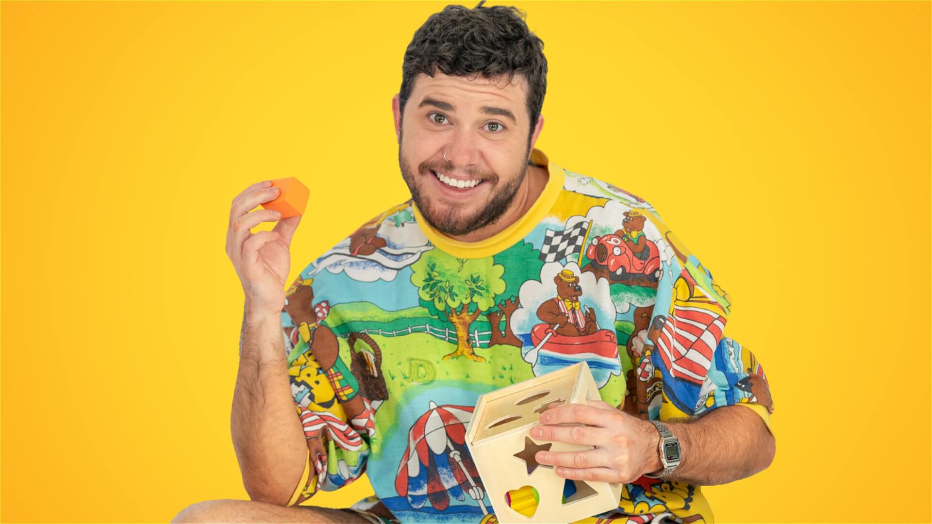 A smiling man in a colorful shirt holds a yellow toy block and a wooden shape-sorting cube against a bright yellow background.