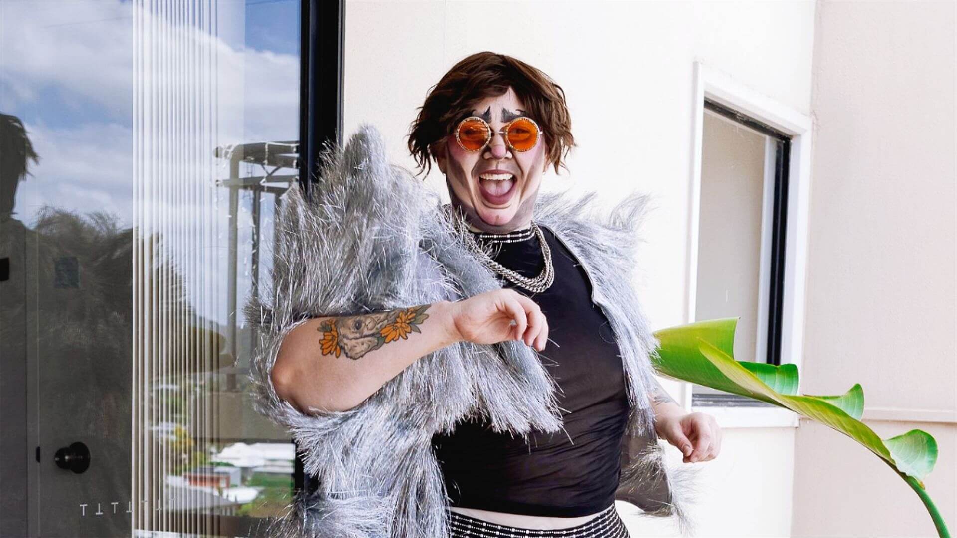 A person wearing round orange sunglasses, a shiny silver fringe jacket, and a black top smiles widely while holding a green leaf. They have a floral tattoo on their forearm and stand in front of a modern glass window.