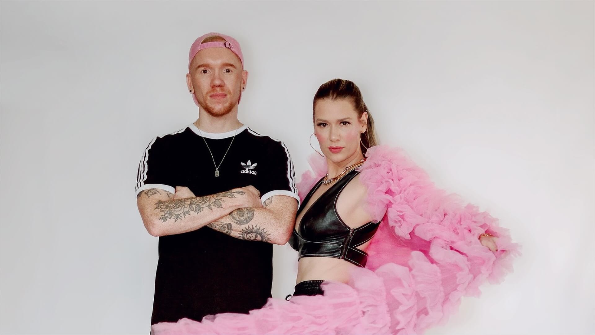 A man in a black Adidas shirt and pink cap stands next to a woman in a black crop top and a dramatic pink ruffled coat. Both look confidently at the camera against a plain white background.