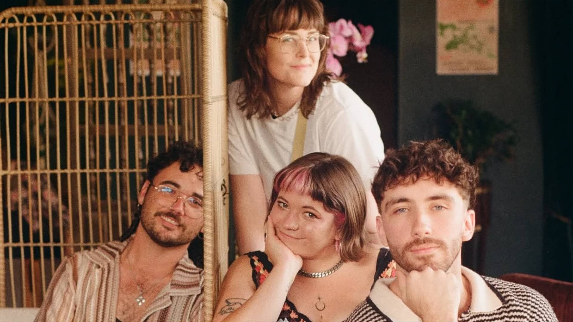 Four young adults, three seated and one standing behind them, pose indoors. They are smiling and relaxed, surrounded by warm lighting and cozy decor, including plants and a wooden screen in the background.