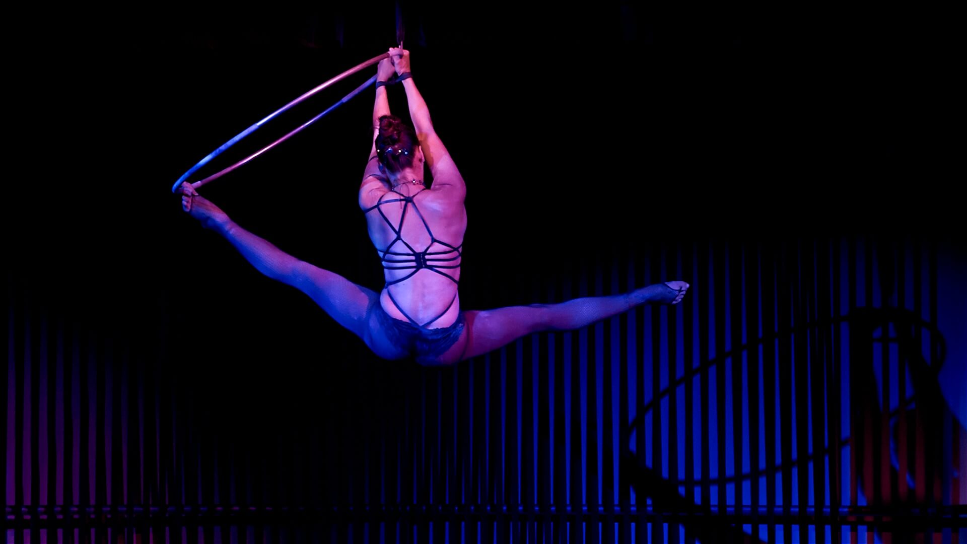 An aerial performer in a strappy costume holds a split position on a suspended hoop, highlighted by dramatic blue and purple stage lighting against a dark, striped background.