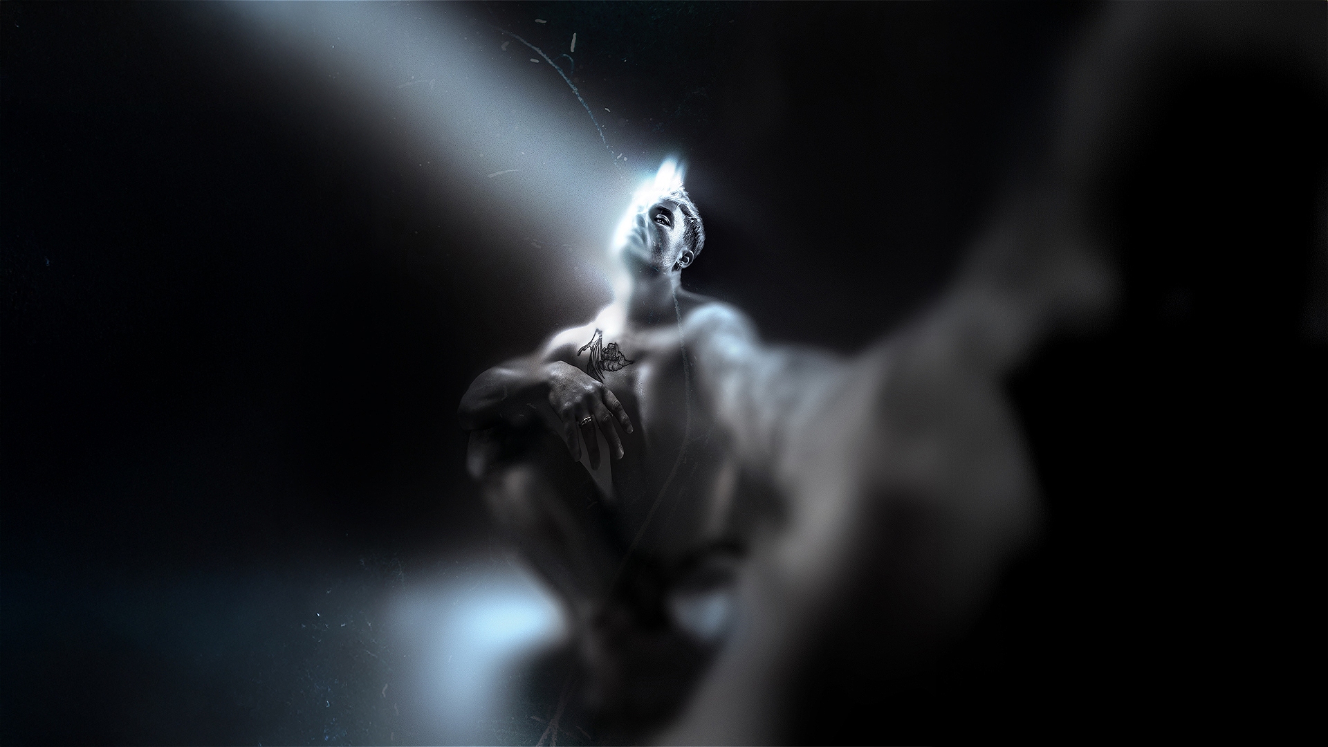 A person sits crouched in darkness, illuminated by a dramatic beam of light from above, creating a surreal, ethereal atmosphere with blurred, glowing edges and a sense of isolation.