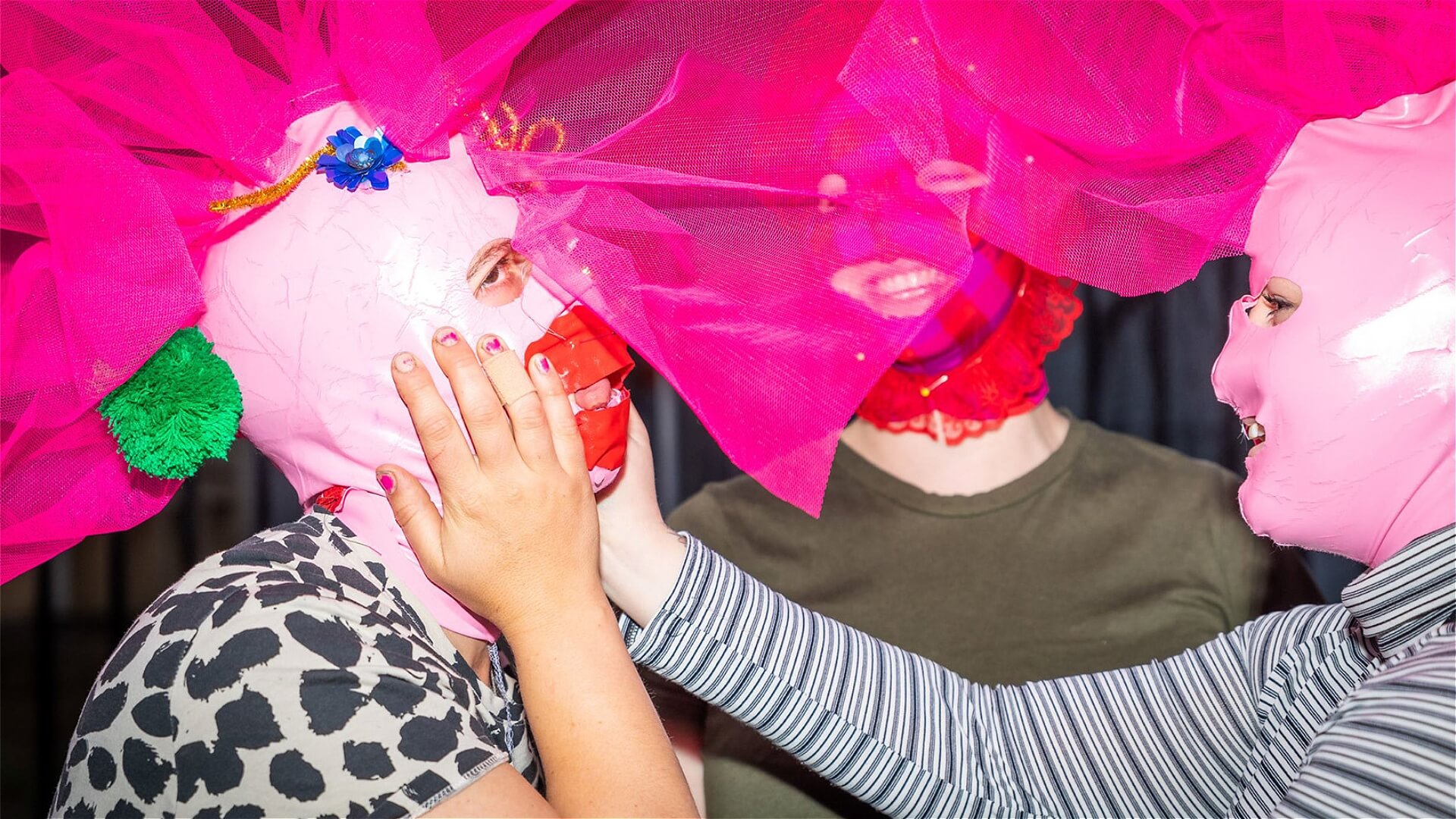 Three people wearing bright pink masks and large, pink tulle headpieces interact playfully. One person touches anothers masked face, and all appear to be enjoying the moment together.