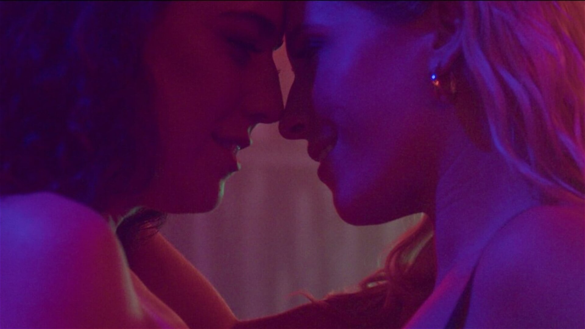 Two women, softly lit with pink and purple lighting, stand very close, gently touching foreheads and smiling intimately, creating a romantic and tender atmosphere.