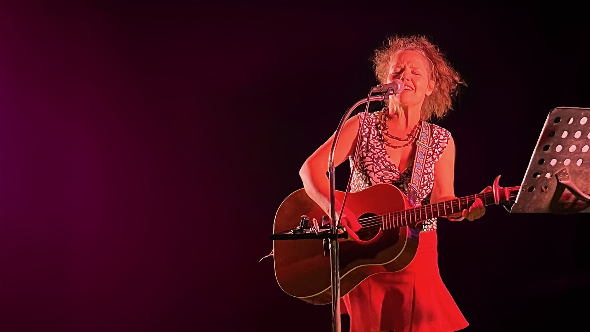 A woman passionately sings and plays an acoustic guitar on stage, standing in front of a microphone. She wears a red skirt and patterned top, with a music stand nearby and a dark, dramatic background.