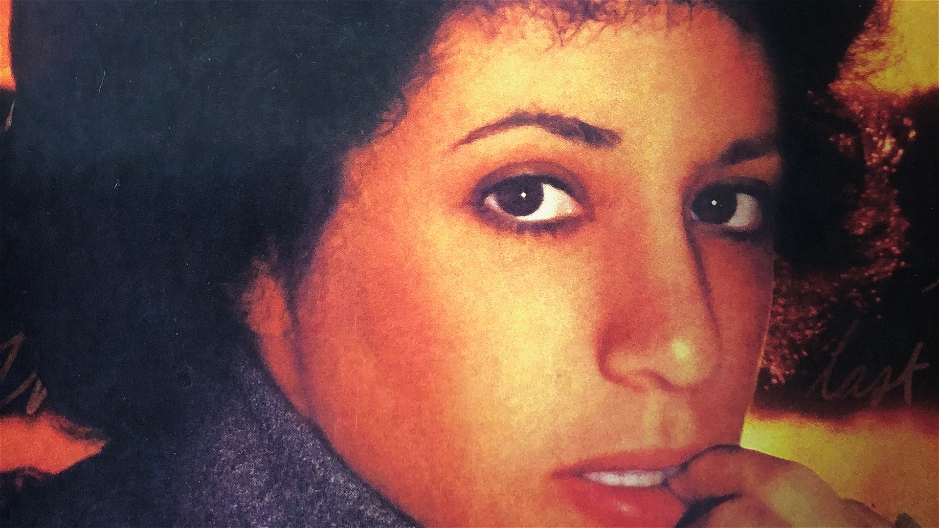Close-up of a person with curly dark hair looking at the camera, hand resting near their mouth, wearing a gray garment, with a warm orange and yellow background.