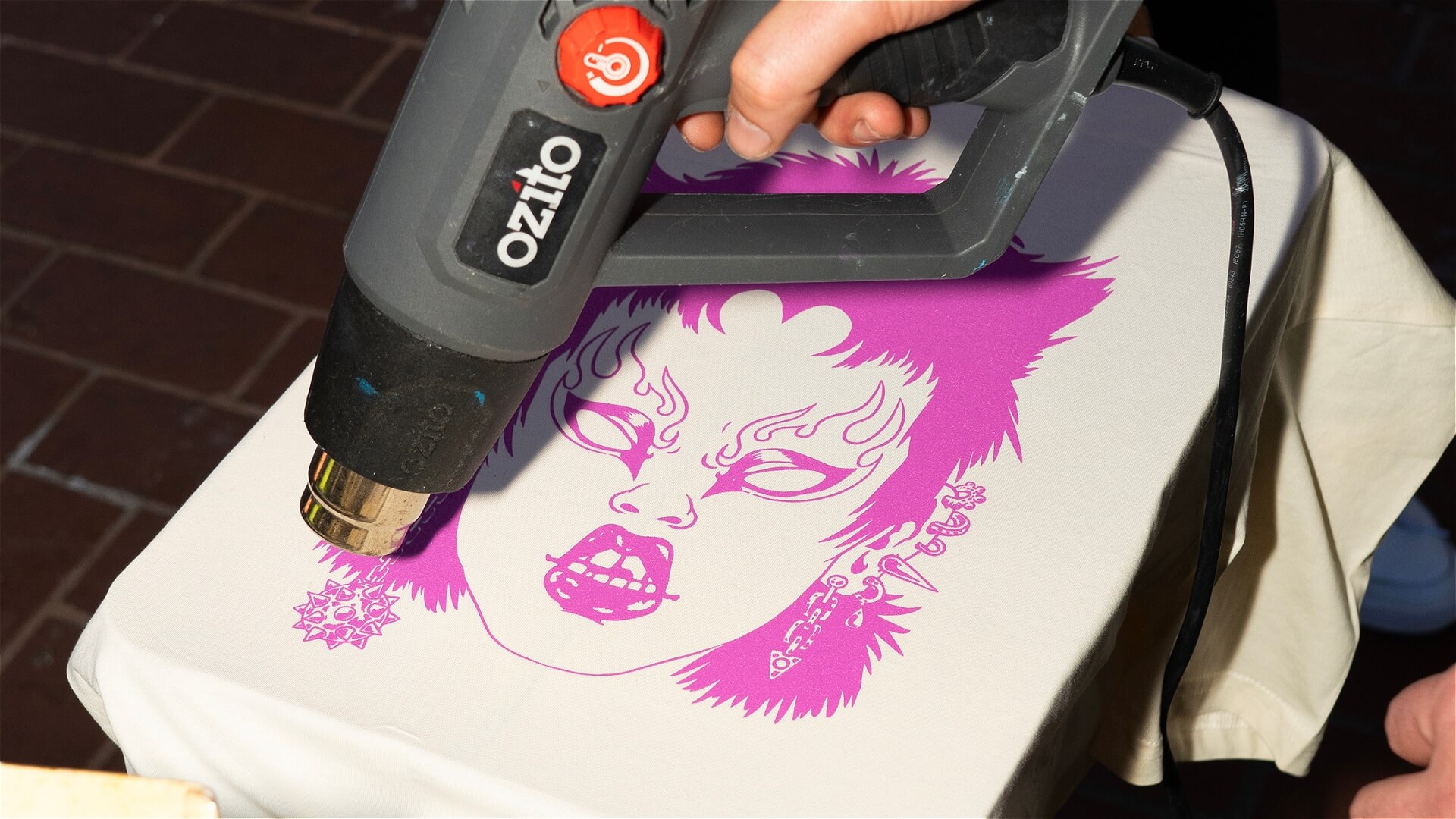 A person uses a black Ozito heat gun on a cream-colored fabric with a bold, purple graphic of a fierce face with fangs, earrings, and dramatic makeup.