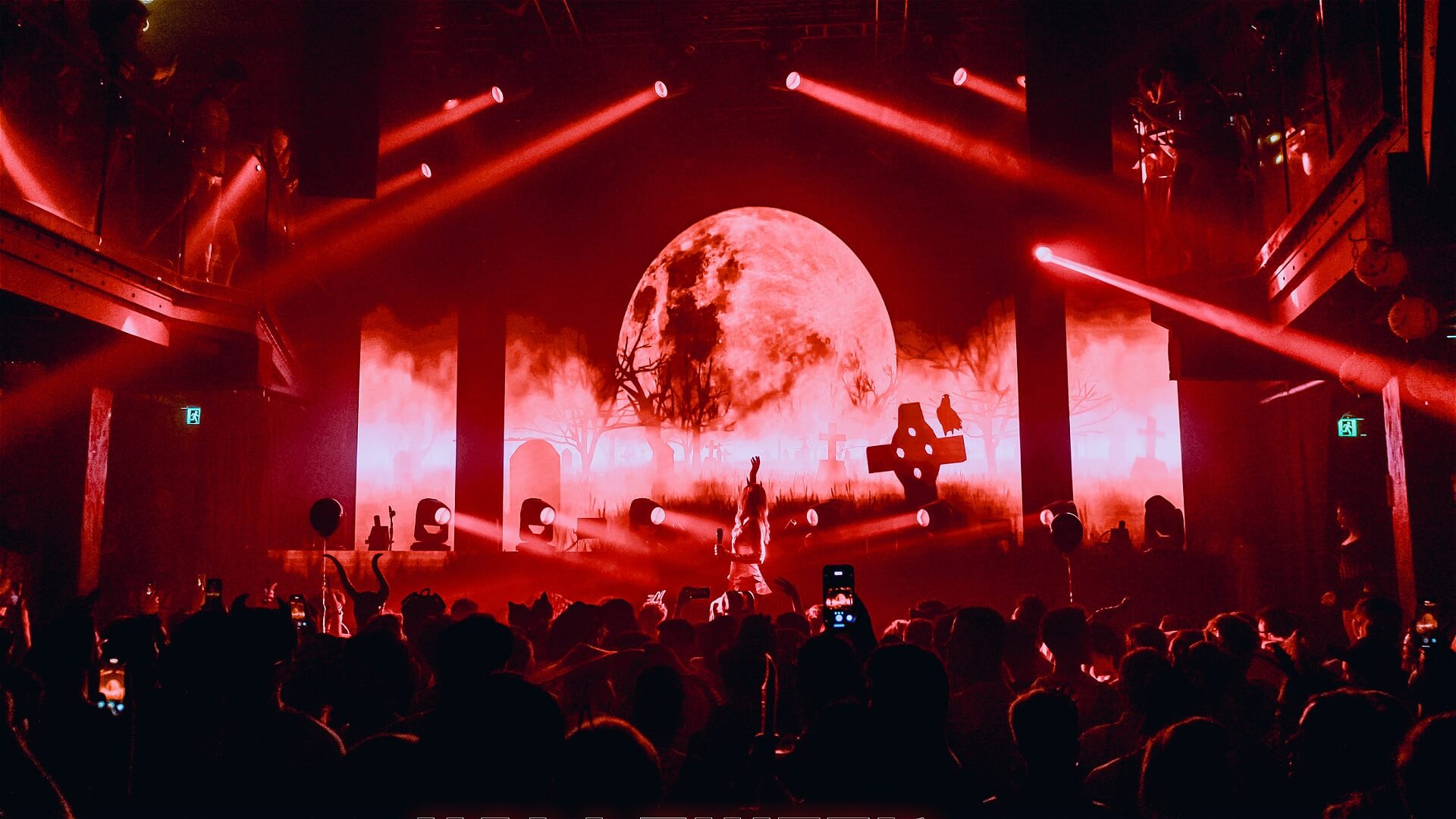 A crowd watches a concert in a dark venue with red lighting, while a large, illuminated full moon and graveyard scene are projected on stage behind the performer.